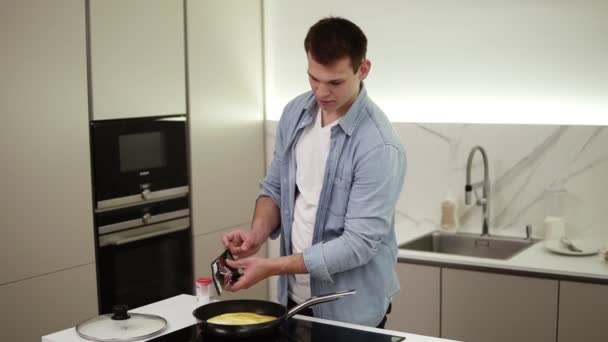 Um homem numa cozinha. Bonito cara alto cozinhando café da manhã, fazendo omelete adicionando especiarias sal, pimenta. Homem alegre de camisa azul gostando de cozinhar. Movimento lento — Vídeo de Stock