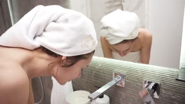 Een vrouw in de badkamer naast de wastafel in witte handdoek op hoofd en lichaam wast haar gezicht met water uit de kraan. Naar de spiegelreflectie kijken en glimlachen — Stockvideo