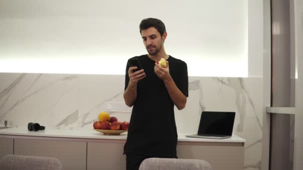 Hombre guapo hojeando en el teléfono móvil en casa cocina luminosa. Guapo joven navegando en el teléfono inteligente sonriendo feliz. Un hombre come una manzana — Vídeo de stock