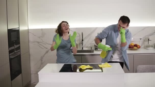 Szczęśliwa młoda para tańcząca w kuchni, zarówno w zielonych gumowych rękawiczkach, bawiąca się w dzień sprzątania w studio apartamencie. Nowoczesna młodzież, ludzie i koncepcja sprzątania — Wideo stockowe