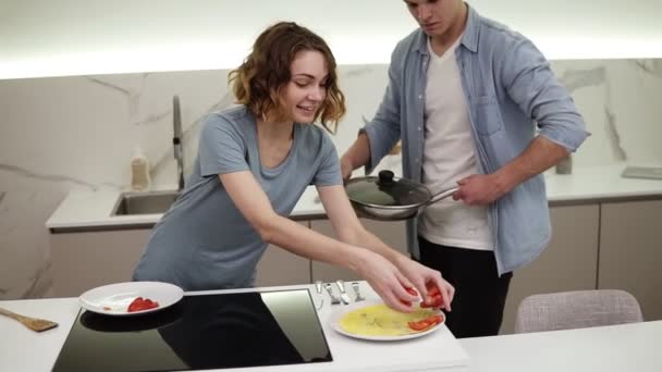 Casal alegre em brilhante, cozinha branca. Eles prepararam omelete na panela, namorada está servindo prato, adicionando tomates. Emocionados e famintos. Movimento lento — Vídeo de Stock