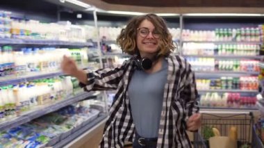 At the supermarket: happy young girl funny dancing between shelves in supermarket. Curly girl wearing jeans and black and white plaid shirt with headphones on neck. Slow motion