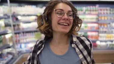 At the supermarket: happy young girl funny dancing between shelves in supermarket. Curly girl wearing black and white plaid shirt with headphones on head listening to her favorite music. Slow motion