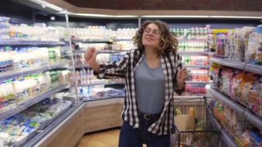 At the supermarket: happy young girl funny dancing between shelves in supermarket. Curly girl wearing jeans and black and white plaid shirt with headphones, listening to the music. Slow motion