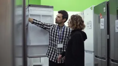 Genç bir kadın, tüketici elektroniği mağazasından almadan önce erkek danışmanla buzdolabının önünde tasarım ve kaliteyi tartışıyor. Depo ile özellikler tartışılıyor