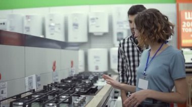 Ekose gömlekli, kulaklıklı bir erkek, mağazada yeni bir fırın seçiyor. Mobilya ve ev aletleri almayı planlıyor, bayan danışmana danışarak ve danışmanlık yaparak.