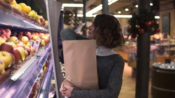 Şirin kız marketten taze kırmızı elmalar alıyor. Güzel genç bir kadın rafın önünde duruyor ve elmaları kahverengi bir kesekağıdına koyuyor, seçiminden memnun. Yavaş çekim — Stok video