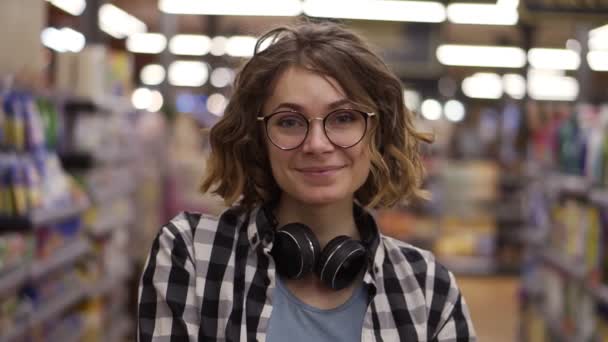 Retrato jovem mulher fica na frente da câmera e sorrisos no supermercado se sentir menina feliz compras loja de varejo rosto. Muito encaracolado cliente feminino em casual com fones de ouvido no pescoço — Vídeo de Stock