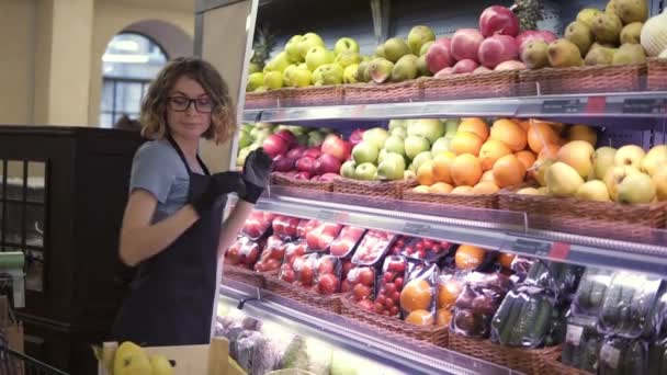 Close up of pretty caucasian worker in black apron and gloves stocking the fruits in supermarket. Young employee in glasses at work. Curly female arranging big red apples on shelf. Slow motion. Side — Stock Video