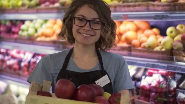 Bonita sorridente jovem funcionária do supermercado feminino em avental preto segurando uma caixa cheia de maçãs na frente da prateleira no supermercado com rosto bonito olhando para a inicialização do retrato frontal profissional da câmera — Vídeo de Stock