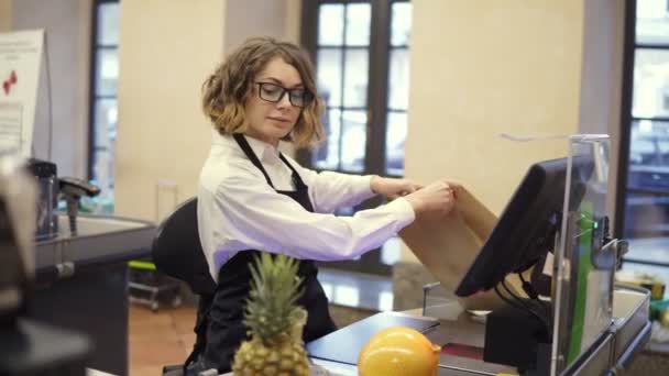 Joyeux vendeur mince en chemise blanche et tablier noir produit de numérisation, fruits au comptoir de caisse dans un supermarché lumineux et le mettre dans un sac en papier brun — Video