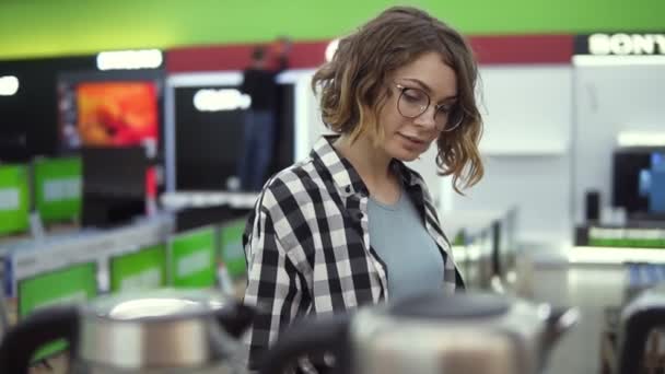 Joven mujer positiva alegre en camisa a cuadros y gafas de elegir hervidor electrónico en la tienda de electrodomésticos, caminando en una fila, sosteniendo y examina la tetera de plata — Vídeos de Stock