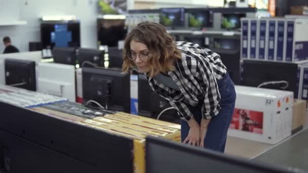 Cliente feminino moderno inteligente escolhendo grandes aparelhos de TV na loja de eletrônicos. Novas gerações de ecrãs. Olhe atentamente em grande tv fina na loja — Vídeo de Stock