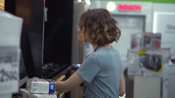 Кудрявая концентрированная женщина-консультант по продажам в бытовом отделе носит и сканирует товары со штрих-кодами в коробках, стоя перед экраном. Медленное движение. Вид сбоку — стоковое видео