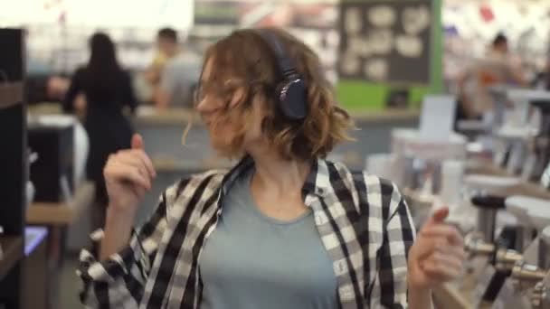 Portret uroczej białej młodej kobiety z krótkimi kręconymi włosami, koszulą w kratę i w słuchawkach tańczącej i słuchającej muzyki w środku supermarketu z domowymi rzeczami. Zwolniony ruch — Wideo stockowe