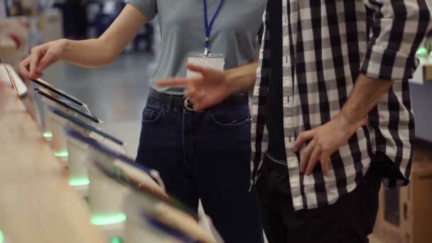 Una consultora irreconocible asesora profesionalmente a un joven con camisa a cuadros. Él considera comprar un nuevo teléfono inteligente y necesita la opinión de expertos. La tienda es moderna, luminosa y tiene todas las novedades — Vídeos de Stock