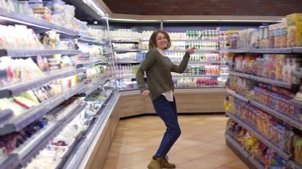 Filmaufnahmen in voller Länge zeigen eine junge Frau in Jeans und Pullover, die am Gang eines Lebensmittelgeschäfts mit großem Sortiment steht. Aufgeregte Frau hat Spaß, tanzt Supermarkt. Zeitlupe — Stockvideo