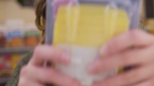Jovem de cabelos encaracolados fazendo compras no supermercado, ela está lendo um rótulo de produto e fatos nutricionais em uma caixa com queijo. Filmagem da prateleira, extremamente close-up do rosto — Vídeo de Stock