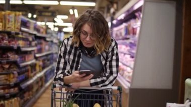 Alışveriş, teknoloji, satış, tüketim ve insan konsepti. Süpermarkette boynunda kulaklık olan kadın. Gülümseyen kadın arkadaşlarıyla mesajlaşıyor, alışveriş arabasına yaslanıyor.