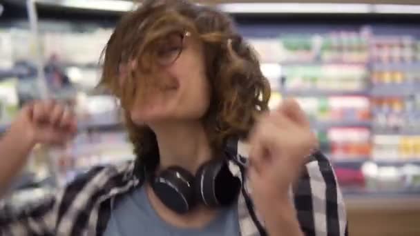 Au supermarché : joyeuse jeune fille drôle de danse entre les étagères dans le supermarché. Fille bouclée portant une chemise à carreaux noir et blanc avec un casque sur le cou. Au ralenti. Gros plan — Video