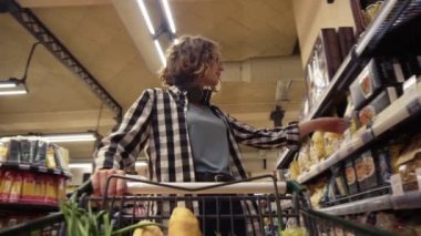 Ekose gömlekli ve kulaklıklı gülümseyen kadın markette, içinde yiyecek olan alışveriş arabasıyla yürüyor. Rafların önünde çeşitli malzemelerle dururken, bir kavanoz al ve el arabasına koy.