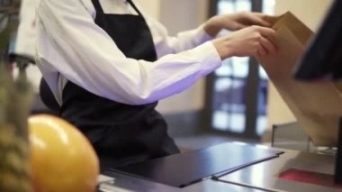 Beyaz gömlekli, siyah önlüklü, tanınmayacak kadar ince bir satış elemanı. Kasadaki meyveler kahverengi kağıt poşete konuyor. Kapat.