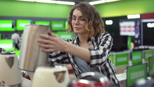 Молодая веселая позитивная женщина в клетчатой рубашке и очках выбирает электронный чайник в магазине бытовой техники, берёт его с полки и осматривает серебряный чайник — стоковое видео