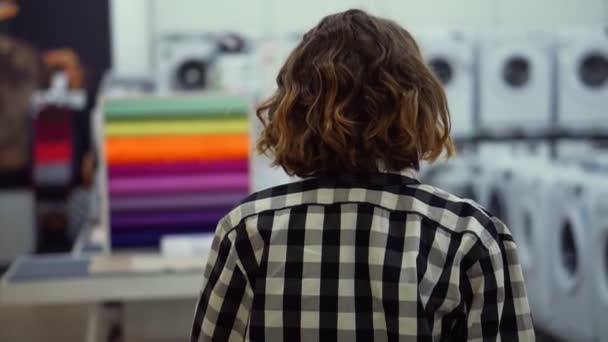 Відстежувати рідкісні зображення дівчини в плетеній сорочці і коротке волосся, що йде по супермаркету в побутовій техніці, шукаючи щось. Розмитий фон — стокове відео