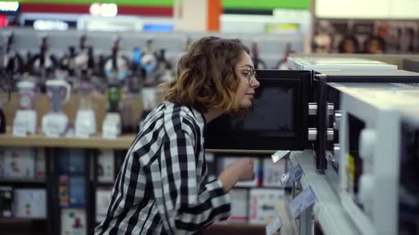 Widok z boku młodej kręconej kobiety w koszuli w kratę wybiera kuchenkę mikrofalową w supermarkecie z elektroniką użytkową. Otwiera drzwi i zagląda do piekarnika. — Wideo stockowe