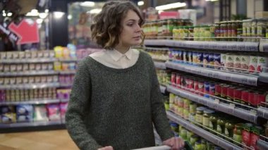 Kafkasyalı kadın market raflarının yanında el arabasıyla markette cam kavanozu kontrol ediyor. Kadın müşteri ürün malzemelerini kontrol ediyor. Süpermarket, satış, alışveriş, çeşitlilik, tüketim