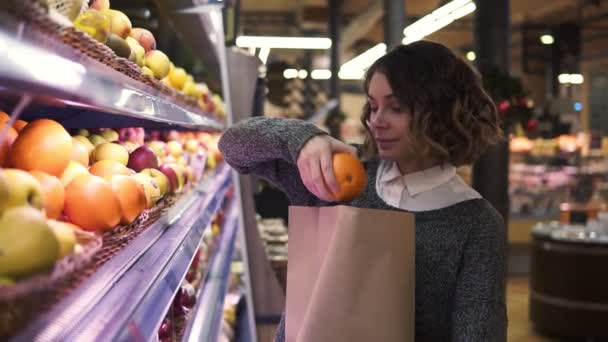 Menina bonito compra laranjas frescas no mercado. Mulher bonita fica na frente da prateleira e coloca as laranjas para um saco de papel marrom, ela está satisfeita com a escolha — Vídeo de Stock