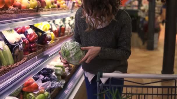 Młoda kobieta we współczesnym supermarkecie wybiera dużą kapustę w dziale warzyw organicznych i umieszcza ją na wózku. Zdrowa kobieta kupująca zielone jedzenie. Koncepcja świeża, asortyment, wegetarianka w spożywczaku — Wideo stockowe