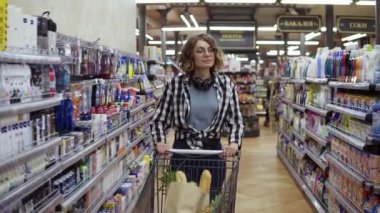 Gözlüklü ve kulaklıklı modern bir genç kız markette alışveriş arabasıyla yürüyor ve etrafta dolanıyor. Ekose gömlekli ve kot pantolonlu kız. Yavaş çekim