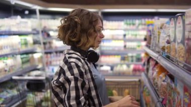 Boynunda kulaklıkla markette yulaf ezmesi ve mısır gevreği seçen genç bir kadının yan görüntüsü. Kahvaltı için mükemmel bir yemek. Etiketleri okumak, en iyi pulları seçmek ve bir arabaya koymak.