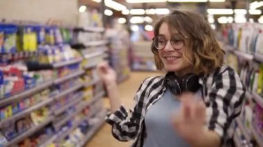 Ekose gömlekli ve gözlüklü genç bir kadının market koridorunda dans ederken çekilmiş portresi. Heyecanlı, boynunda kulaklıkla gülümseyen, eğlenen, süpermarkette dans eden bir kadın. Yavaş çekim