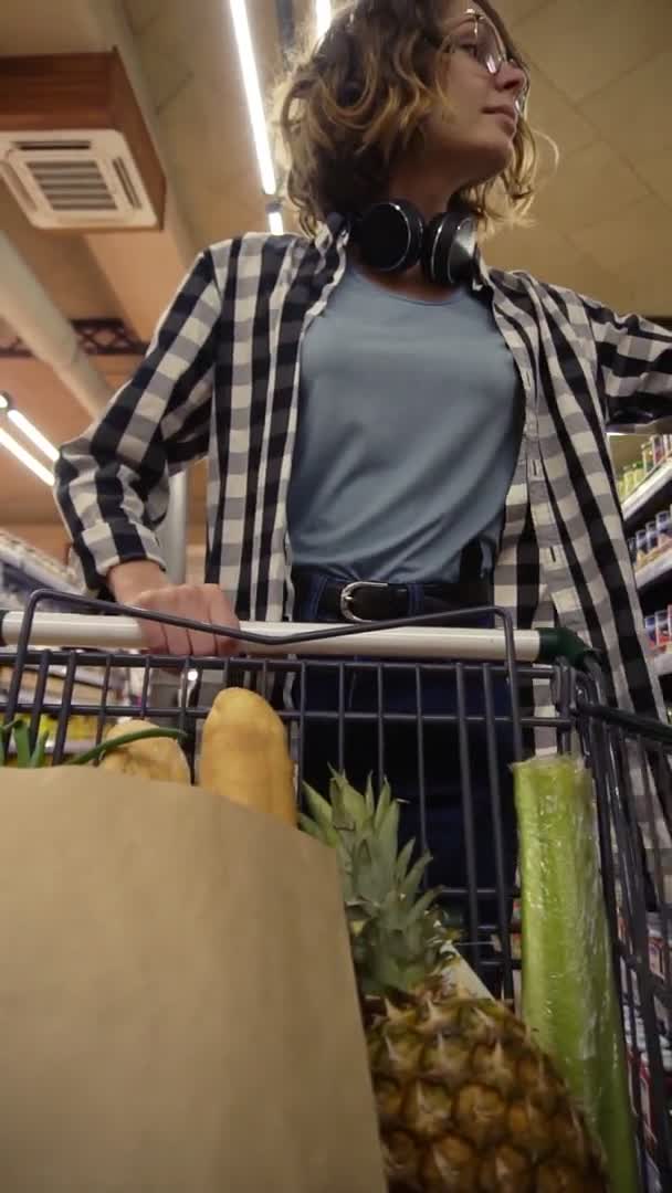 Leende kvinna i rutig skjorta och hörlurar på halsen går i mataffären styr shoppingvagn med mat inuti och titta runt på hyllor med produkter, ta en burk och sätta den till en — Stockvideo
