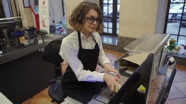 Fröhliche, schlanke Verkäuferin in weißem Hemd und schwarzer Schürze scannt Produkt, Obst an der Kasse im hellen Supermarkt und steckt es in braune Papiertüten. Filmmaterial aus dem Hochwinkel — Stockvideo