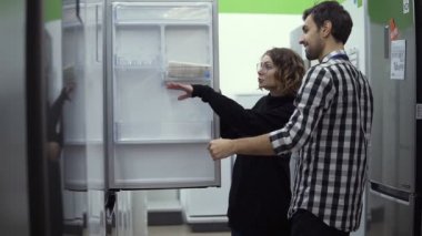 Genç evli çift, tüketici elektroniği mağazasından almadan önce açık buzdolabını, tasarımı ve kalitesini inceliyor. Özellikler ve tasarım tartışılıyor