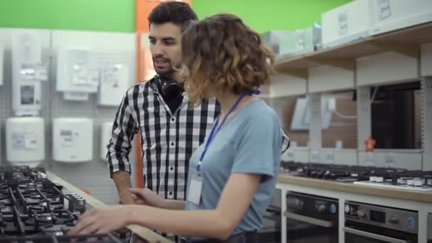 Hombre moreno y sonriente en camisa a cuadros con auriculares eligiendo un nuevo horno moderno en la tienda. Está planeando comprar muebles y electrodomésticos, chequeando y asesorando con una consultora — Vídeos de Stock