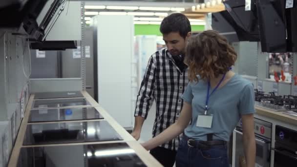 Hombre moreno y sonriente en camisa a cuadros con auriculares eligiendo un nuevo top cocinero moderno en la tienda. Él está planeando comprar muebles y electrodomésticos, consultar y asesorar a la consultora sobre — Vídeo de stock