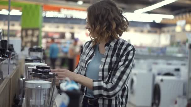 Nel negozio di elettrodomestici, una donna riccia bruna con una camicia a quadri sceglie un frullatore per lo shopping visualizzando e tenendo in mano il top del dispositivo. Vista laterale — Video Stock