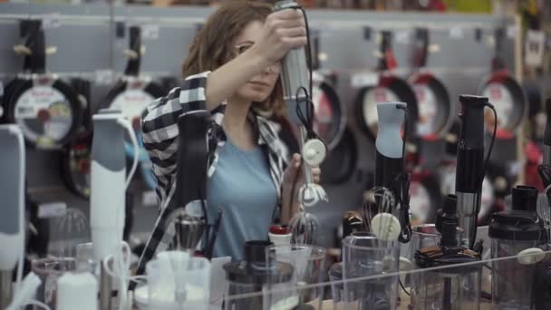 En la tienda de electrodomésticos, una atractiva mujer rizada con una camisa a cuadros elige un palo de licuadora al ver y sostener el dispositivo en sus manos. Movimiento lento — Vídeos de Stock