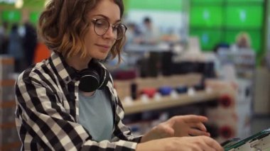 Boynunda kulaklıkları olan kıvırcık bir kadın cep telefonlarıyla tezgahta duruyor ve modern bir elektronik mağazasında yeni bir akıllı telefon seçiyor.