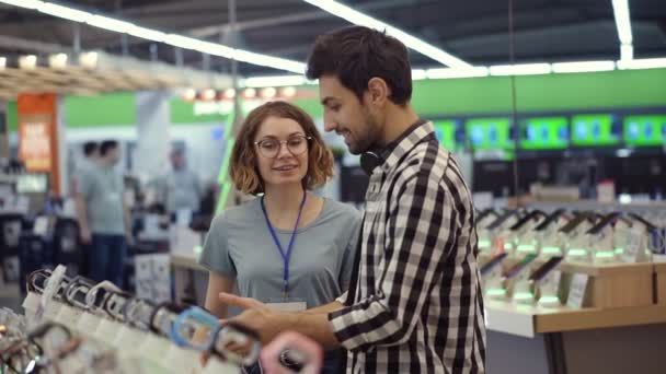 In the electronics store female consultant gives professional advice to a young man. He considers to buy a new smartphone and needs expert opinion. Store is modern, bright and has all the new devices — Stock Video