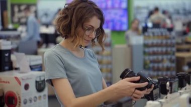 Gözlüklü genç bir kadın elektronik mağazasından yeni bir fotoğraf makinesi alıyor. En iyi modele karar vermeye çalışıyorum. Şüphelerim var. Bir kamerayı kontrol ediyorum.