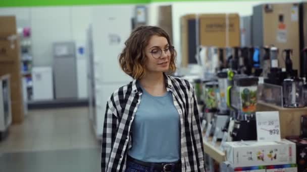 Rastreando imagens da visão frontal de uma menina em uma camisa xadrez e cabelo encaracolado curto andando pelo supermercado em eletrodomésticos à procura de algo. Movimento lento — Vídeo de Stock