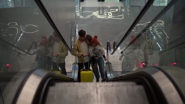 Una familia joven y feliz con una maleta amarilla subiendo por la escalera mecánica desde la sala del aeropuerto. La madre sonriente tiene una hija bonita. Felices abrazos pareja, primeras vacaciones juntos — Vídeo de stock