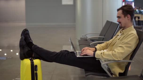 Un jeune homme barbu est assis à l'aéroport, attendant le départ de l'avion et utilisant un ordinateur portable. Avoir une conversation vidéo via une caméra web, tenant les jambes sur sa valise jaune. Mouvement lent — Video