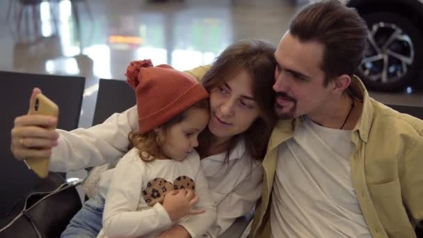Ung familj gör en selfie med smart telefon i väntan på flygplan. Familjen väntar på flyget på internationella flygplatsen. Mamma håller i smarttelefonen och lilla dottern trycker på knappen — Stockvideo