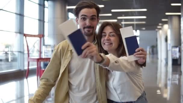 Retrato de casal caucasiano animado saindo de férias. Passando pelo aeroporto e segurando passaportes, cartões de embarque. Acenando com passaportes de check-in, sorrindo para a câmera, abraçando — Vídeo de Stock
